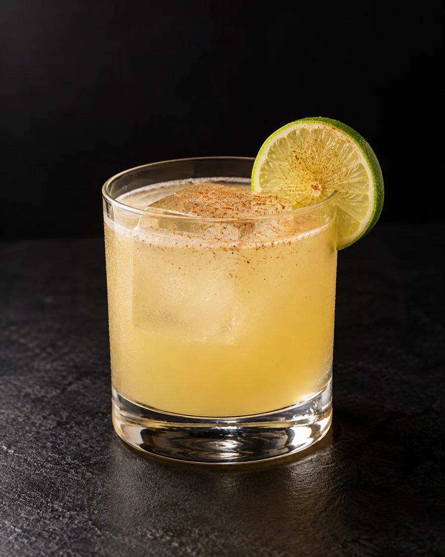 pale cloudy yellow Chitty Chitty Bang Bang over one large cube in a rocks glass with lime wheel and cinnamon dusting.