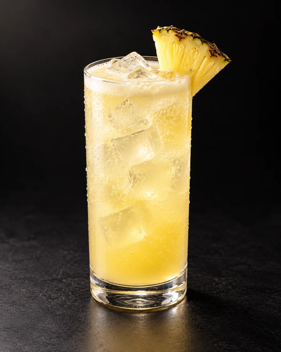 Pineapple Coconut Fizz served in a highball glass with a pineapple wedge against a black background.